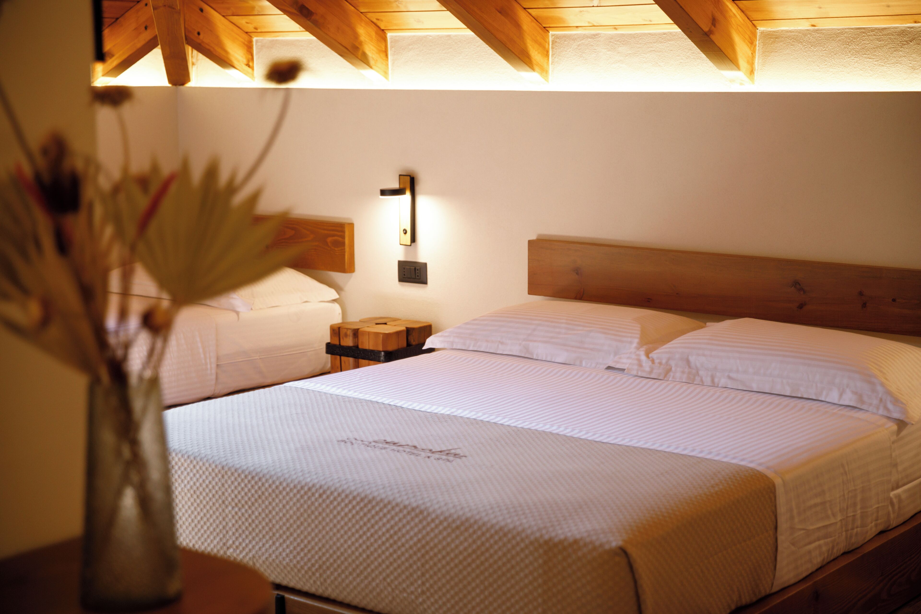 A bedroom with a wooden ceiling, a bed with white bedding, a bedside table with a lamp, and a vase with flowers.