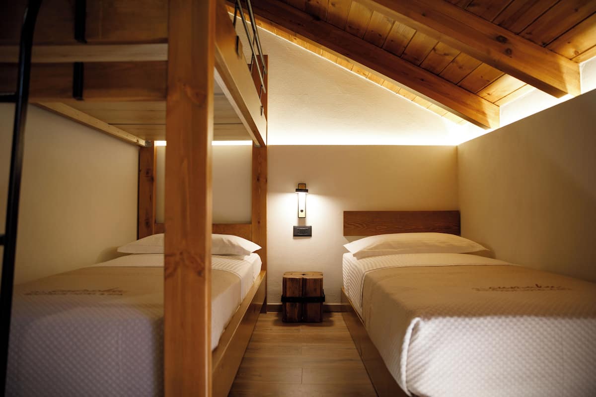 A room with two beds, wooden beams, a small wooden stool, and a wall-mounted light fixture.