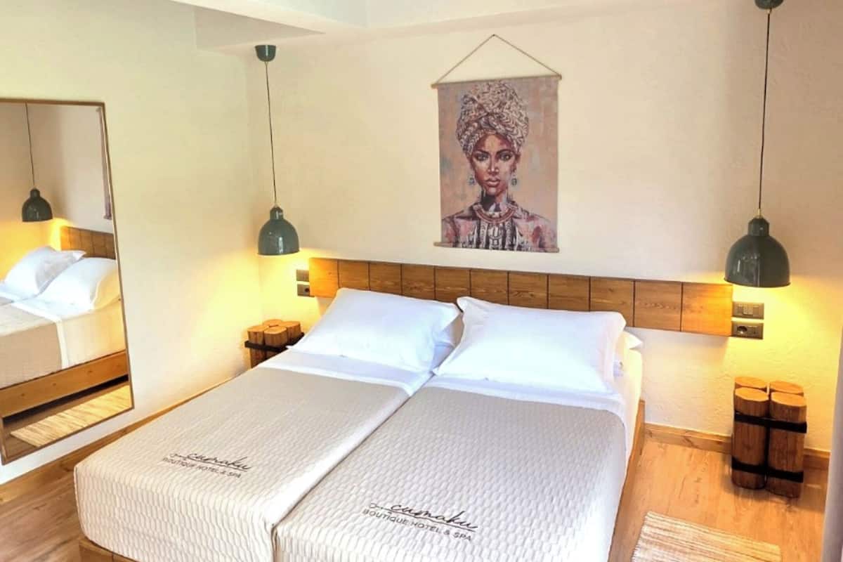A double bed with a headboard, two bedside tables, a wooden floor, a painting on the wall, and pendant lights.