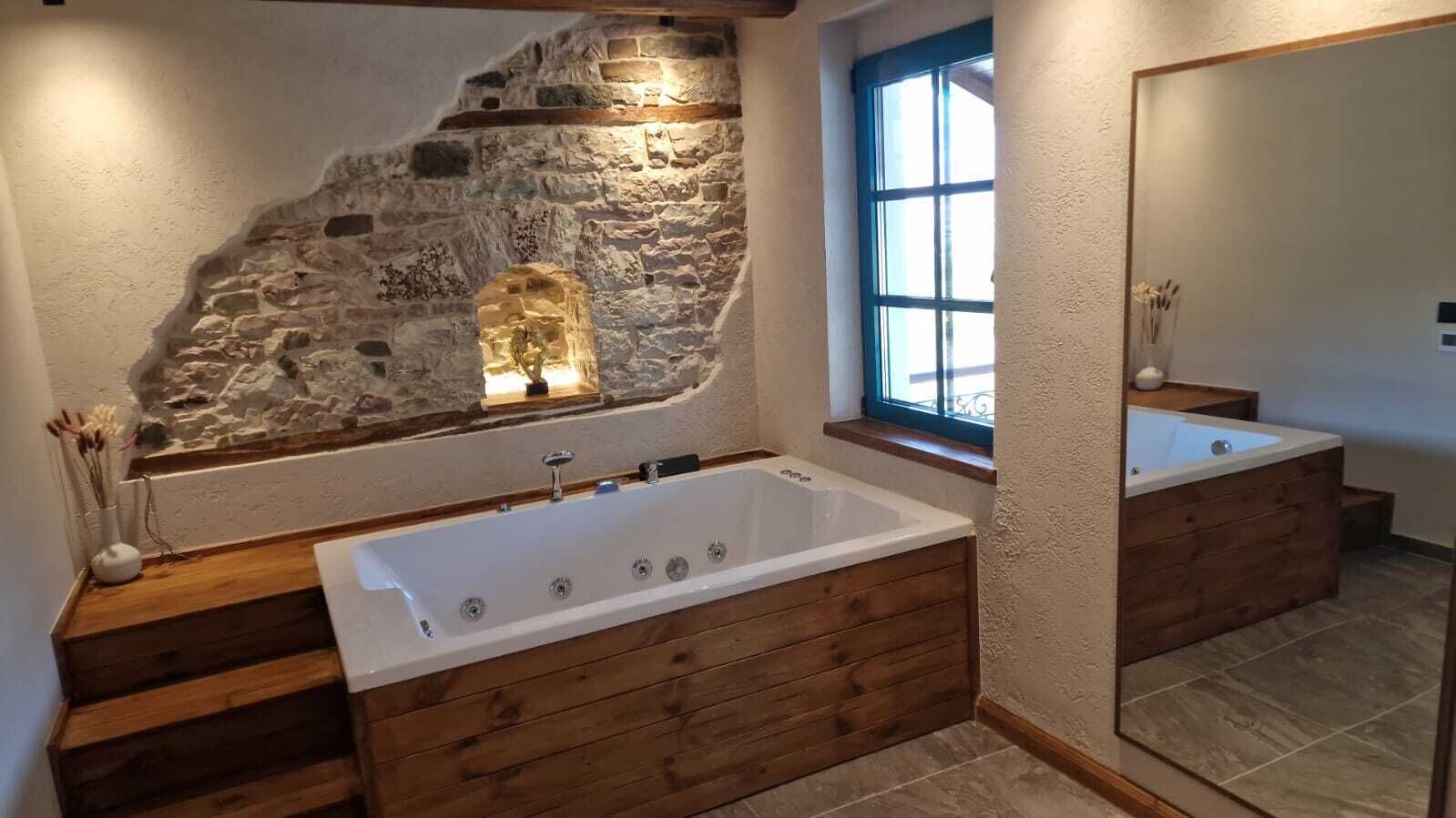Une salle de bain avec un mur en pierre, une grande baignoire, des marches en bois et une fenêtre avec des bordures bleues.