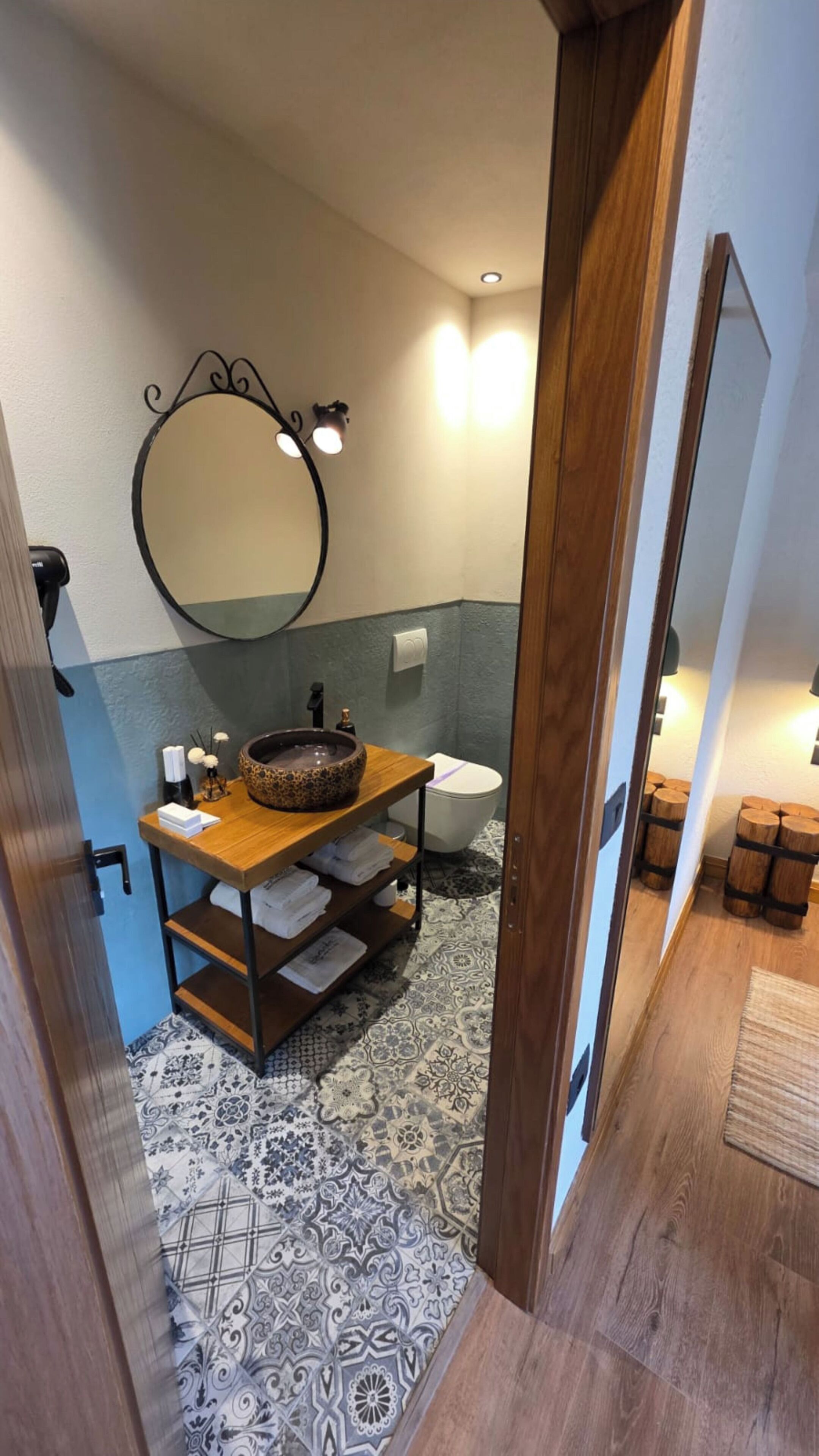 Een badkamer met een houten wastafelmeubel, een ronde spiegel, een toilet en vloertegels met een patroon.