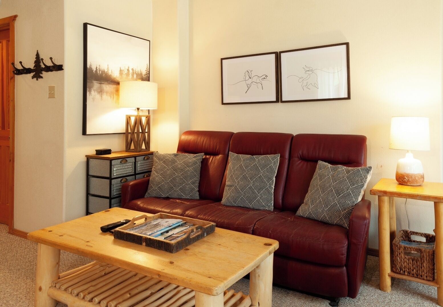 Un salon avec un canapé en cuir marron, deux coussins décoratifs à motifs, une table basse en bois sur laquelle est posée une pile de magazines, une table d’appoint avec une lampe, et des œuvres d’art encadrées accrochées au mur.