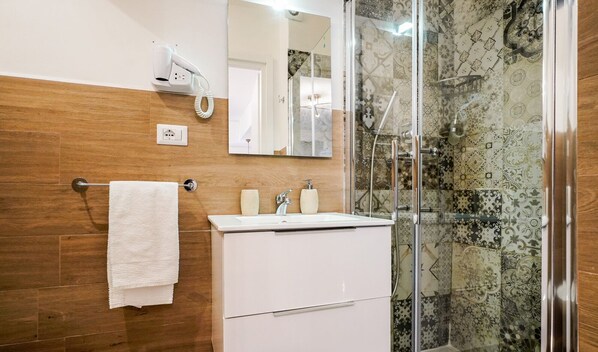 A bathroom with a wooden wall, a white sink, a mirror, a towel rack, and a shower with patterned tiles. - Barirooms - Urban 163 (Bari)