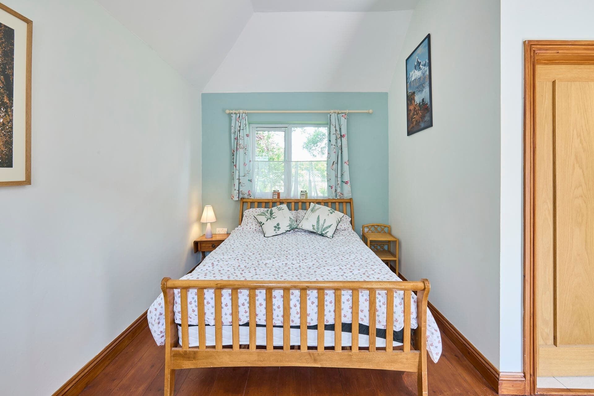 Un dormitorio con cama de madera, una ventana con cortinas florales, una lámpara de noche y un cuadro en la pared.