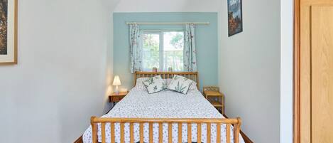 Un dormitorio con cama de madera, una ventana con cortinas florales, una lámpara de noche y un cuadro en la pared.