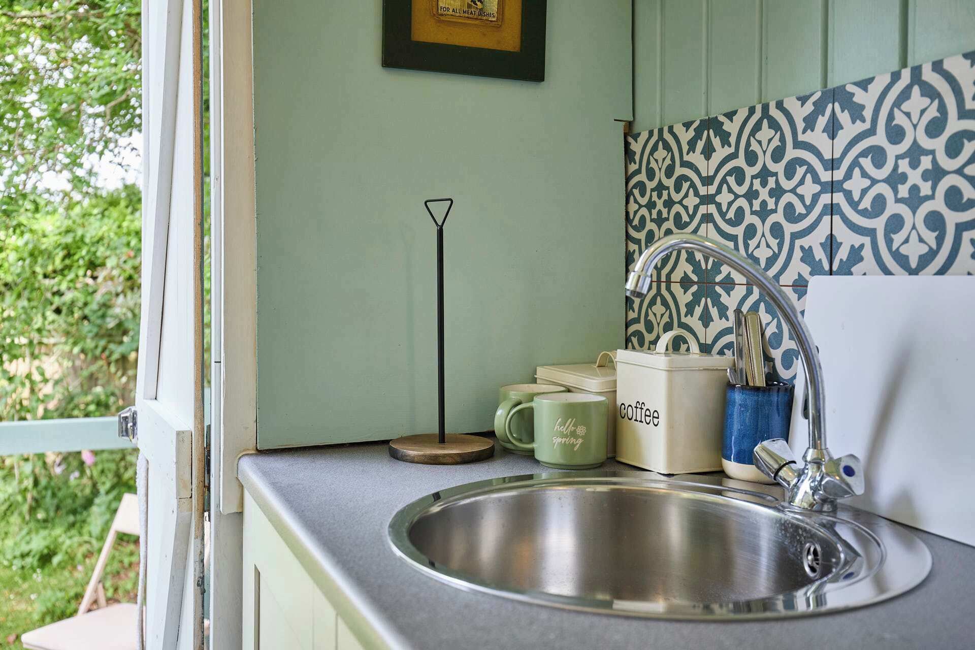 A kitchen with a stainless steel sink, a green mug with 'hello spring' text, a coffee maker, and a patterned backsplash.