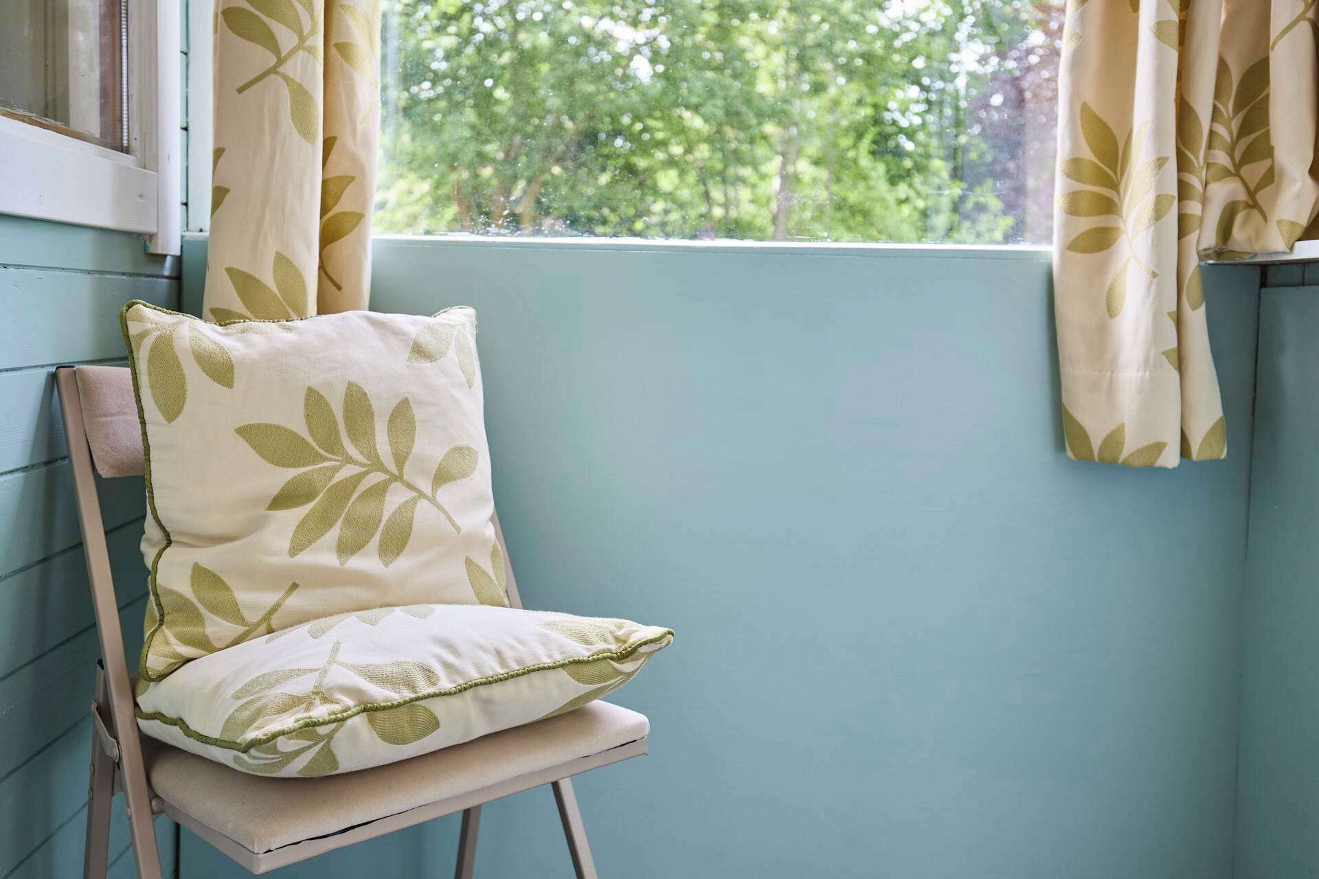A chair with a cushion against a wall, a window with curtains, and a view of trees outside.