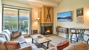 A cozy living room with a fireplace, a sofa, a coffee table, and a dining table. There is a large window with a view of the mountains. - Trails End Après Penthouse Condo - Ski In/Out, Walk Downtown! (Breckenridge)