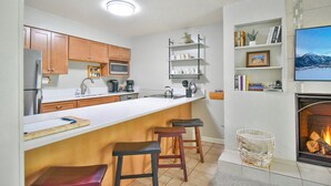 Una cocina moderna con armarios de madera, encimera blanca y microondas integrado. Hay tres taburetes en la isla y una chimenea con un televisor montado encima.