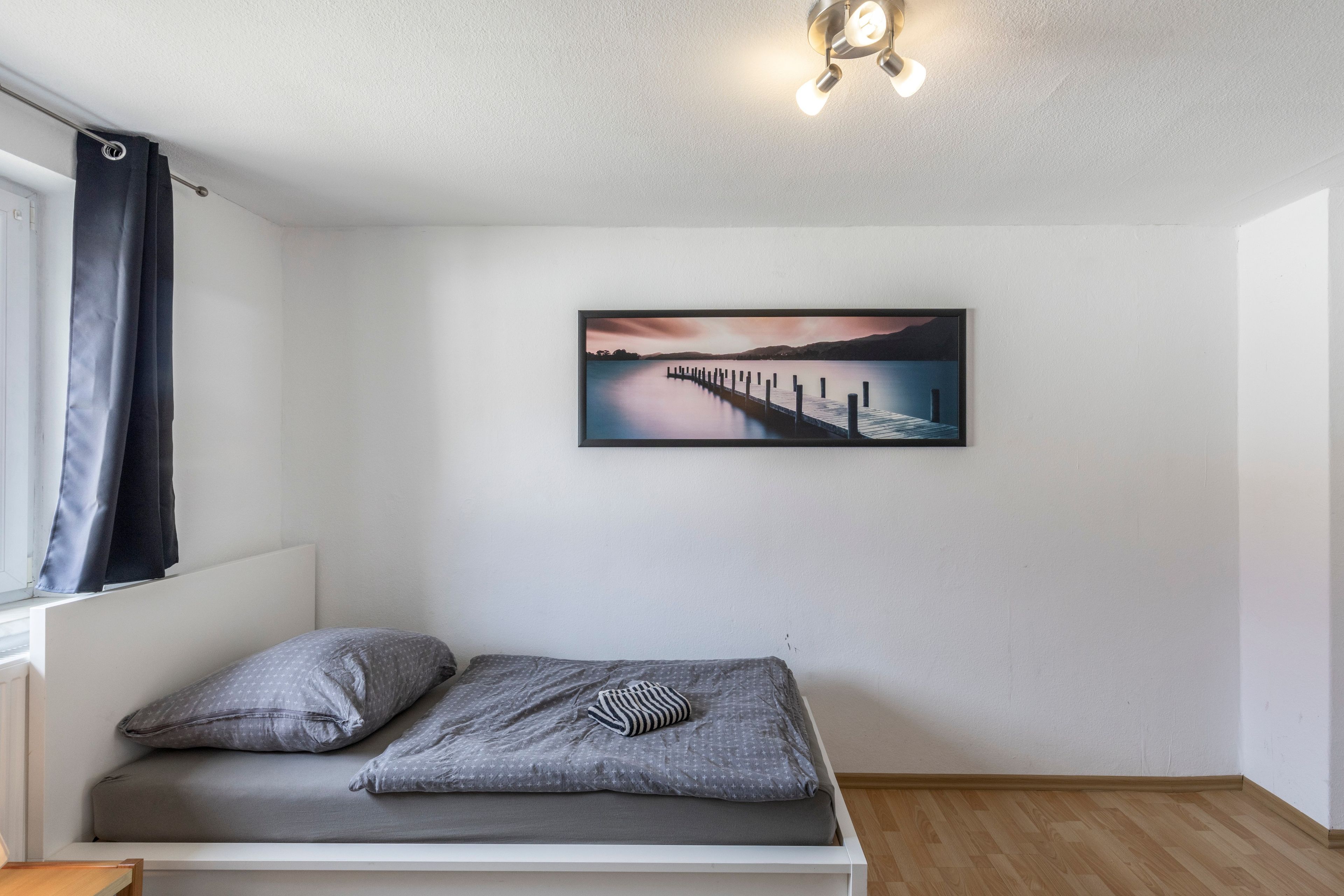 A single bed with a gray mattress and pillows, a framed landscape painting on the wall, and a ceiling light fixture.