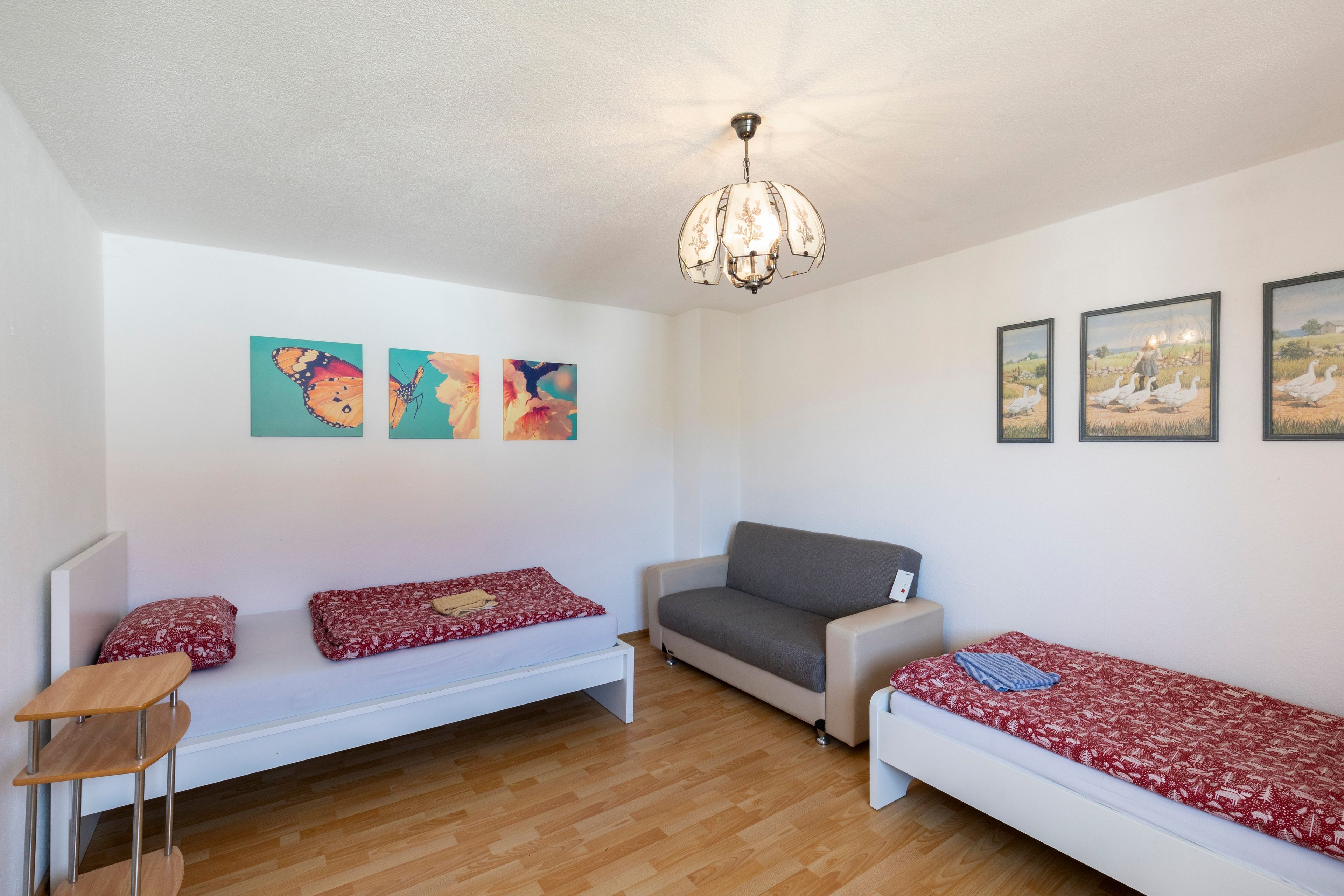 Une petite pièce bien éclairée, avec un plancher en bois, un canapé blanc, un fauteuil gris et une petite table. Trois œuvres d’art encadrées ornent le mur, et un luminaire est fixé au plafond.