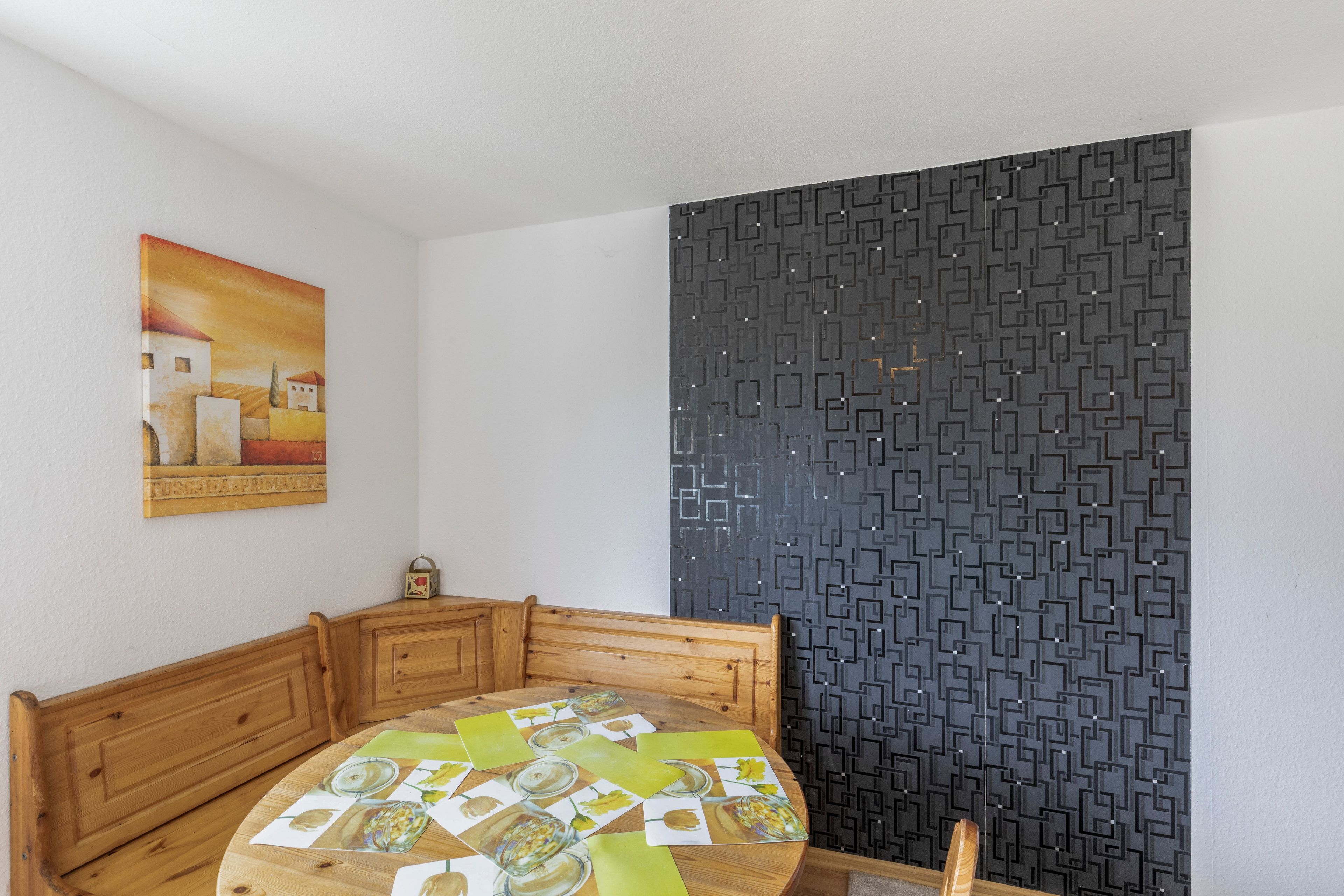 Ein Zimmer mit einer hölzernen Bank, einem Tisch mit kariertem Tischläufer, einer Wand mit geometrischem Muster und einem Gemälde an der Wand.