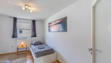 Une chambre à coucher avec un lit, une table de chevet avec une lampe, une fenêtre avec des rideaux, un radiateur et un tableau au mur.