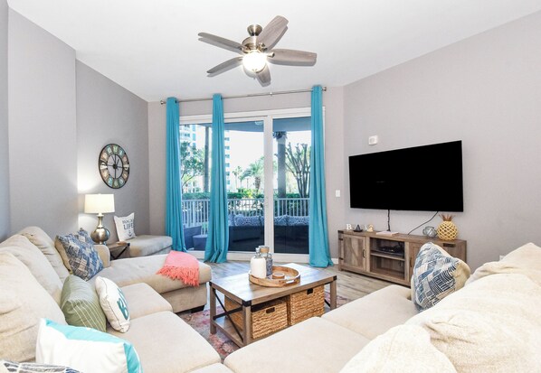 A living room with a ceiling fan, a flat-screen TV, a sofa set, a coffee table, and a window with curtains.