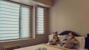 A hotel room with a bed, two windows with blinds, a vent, and a rolled towel on the bed. - One Palm Tree Villas Condo (Pasay)