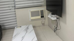 A room with a marble-topped table, a wall-mounted TV, a window with blinds, and a built-in air conditioning unit. - One Palm Tree Villas Condo (Pasay)