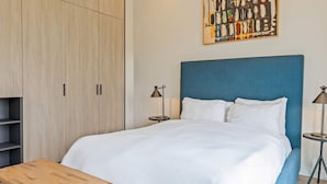 Una cama bien hecha con cabecera azul, una mesita de noche de madera con lámpara y un armario con acabado en madera clara.