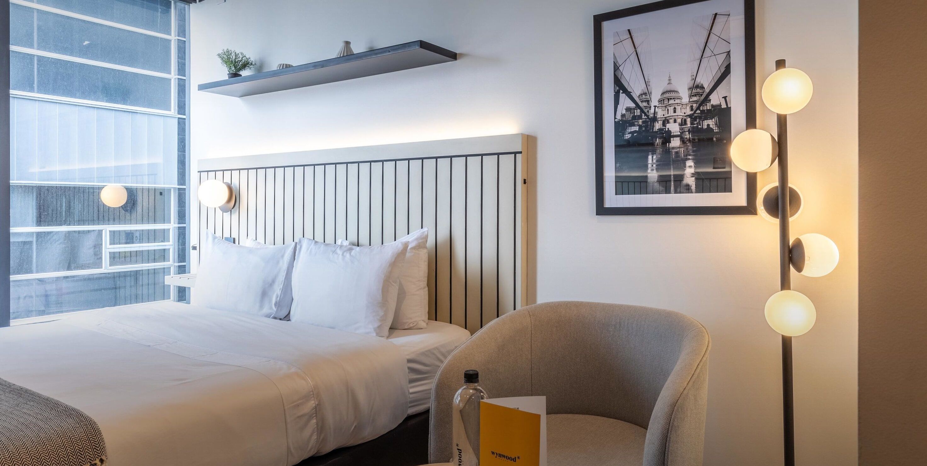 A hotel room with a bed, a chair, a bedside table, a framed picture on the wall, and a shelf with decorative items.