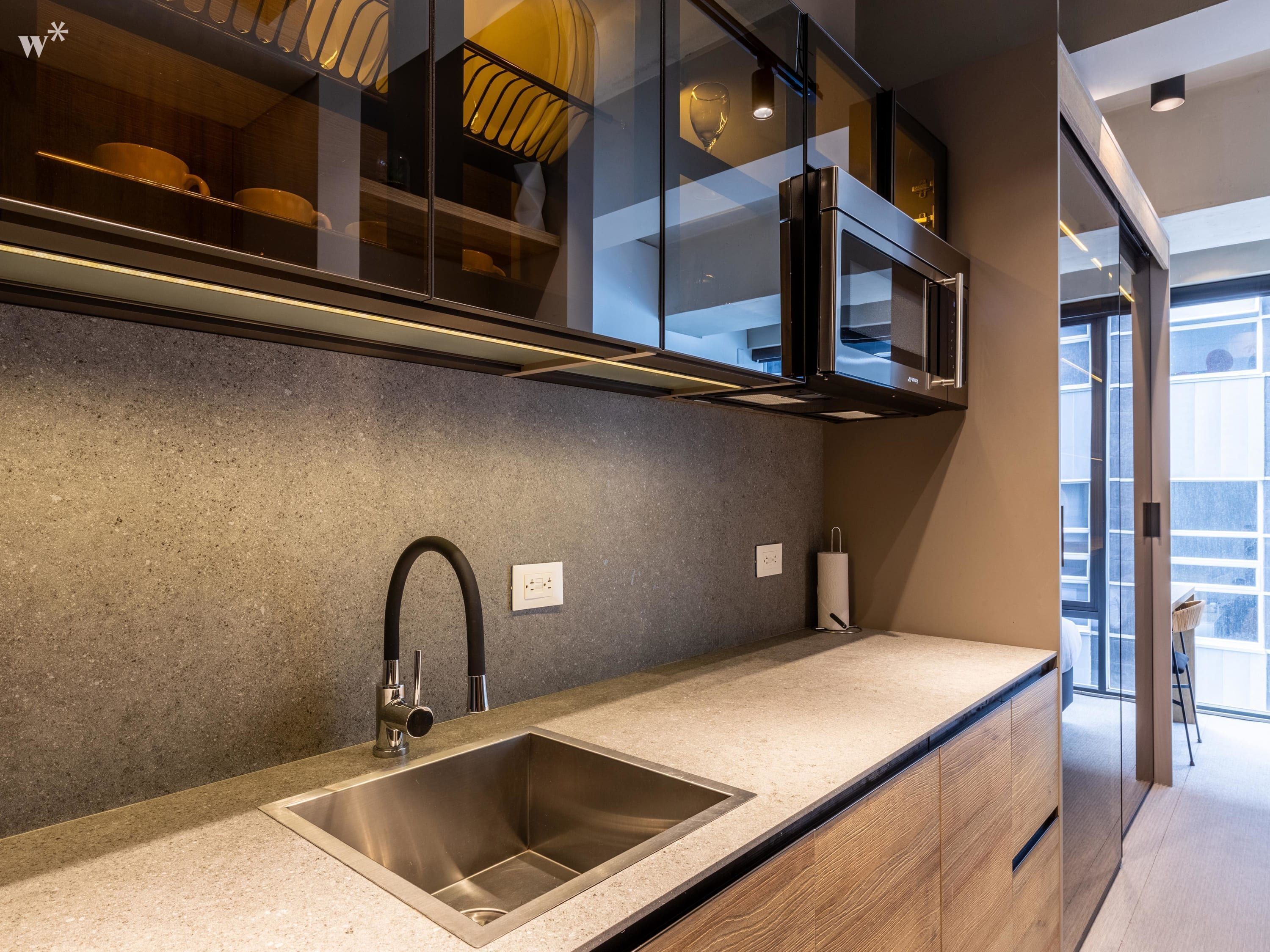 A modern kitchen with a stainless steel sink, wooden cabinets, and a glass-fronted upper shelf with dishes.