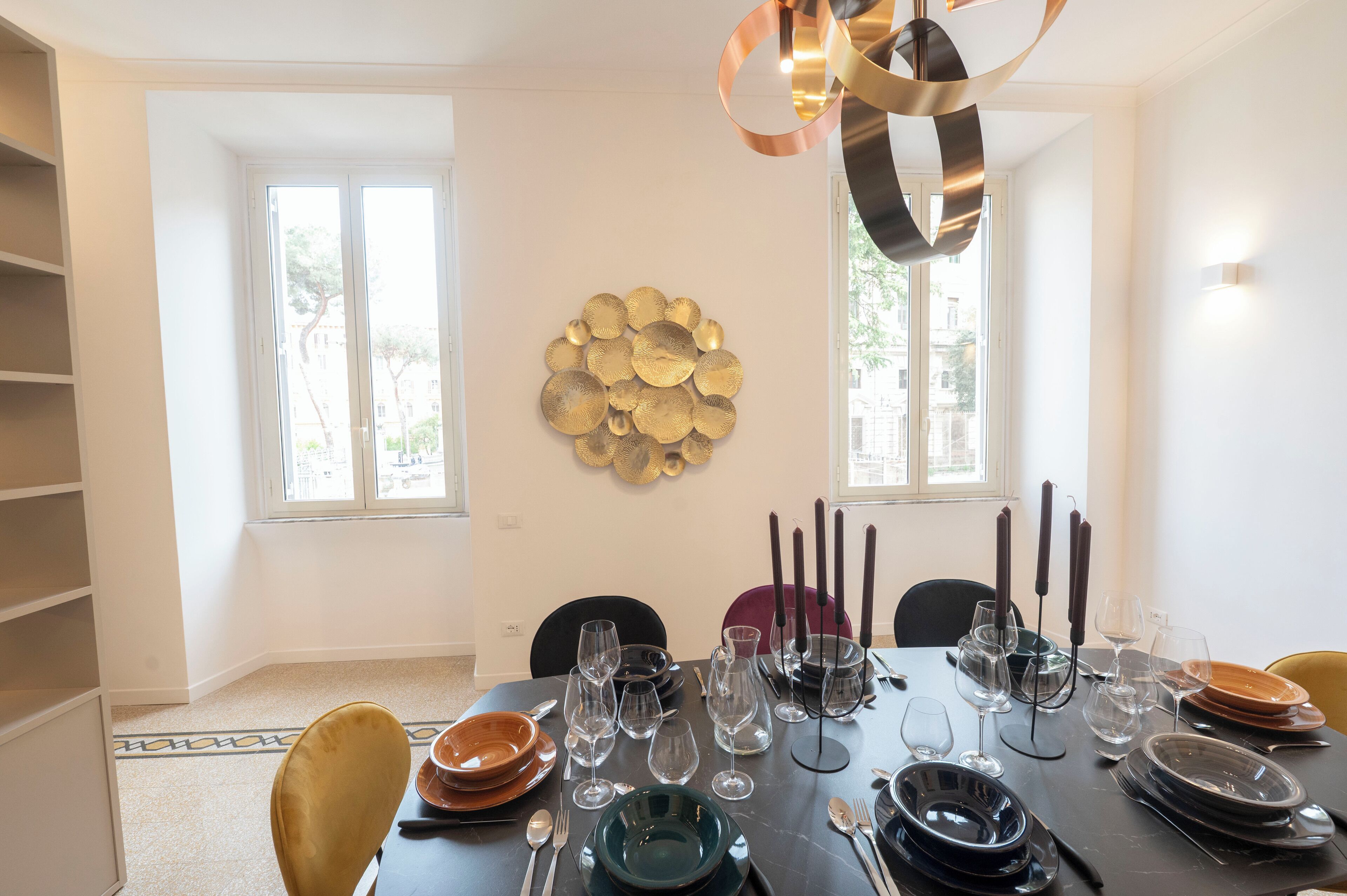A dining area with a table set for six, including plates, glasses, and cutlery. There are two windows providing natural light, and a decorative wall piece above the table.