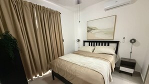 A bedroom with a bed, curtains, a bedside table, a nightstand, a wall-mounted air conditioner, and a painting. - NOK Perfect 1 BR by Vistacana Rentals (Punta Cana)