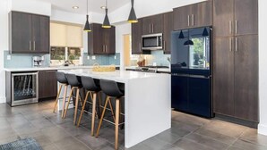 A modern kitchen with dark wood cabinets, a white island with bar stools, a built-in microwave, and a refrigerator. - Gated Home With Pool Hot Tub (Vista)