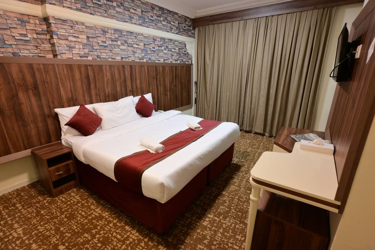 Habitación de hotel con una cama grande, cabecero de madera, mesita de noche pequeña de madera y una mesilla con una caja de pañuelos, y pared con diseño de ladrillo.
