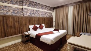 Quarto de hotel com parede de padrão em pedra, cabeceira de madeira, cama com lençóis brancos e almofadas vermelhas, mesa de cabeceira com telefone e escrivaninha com abajur.