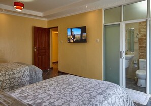 Habitación de hotel con dos camas, un televisor montado en la pared y un baño al que se accede por un cristalero corredizo.