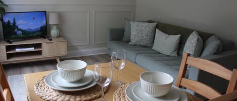 Une table à manger dressée avec des bols blancs et des verres à vin, une télévision et un canapé en arrière-plan.