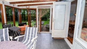 Ein überdachter Patio-Bereich mit Tisch und zwei gestreiften Stühlen, eine Tür zu einem Essbereich und Blick auf den Garten.