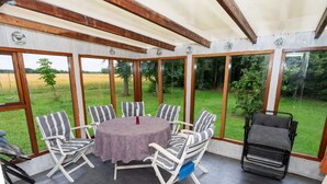 Eine überdachte Terrasse mit einem runden Tisch und sechs gestreiften Stühlen, große Fenster mit Blick auf eine Wiese.