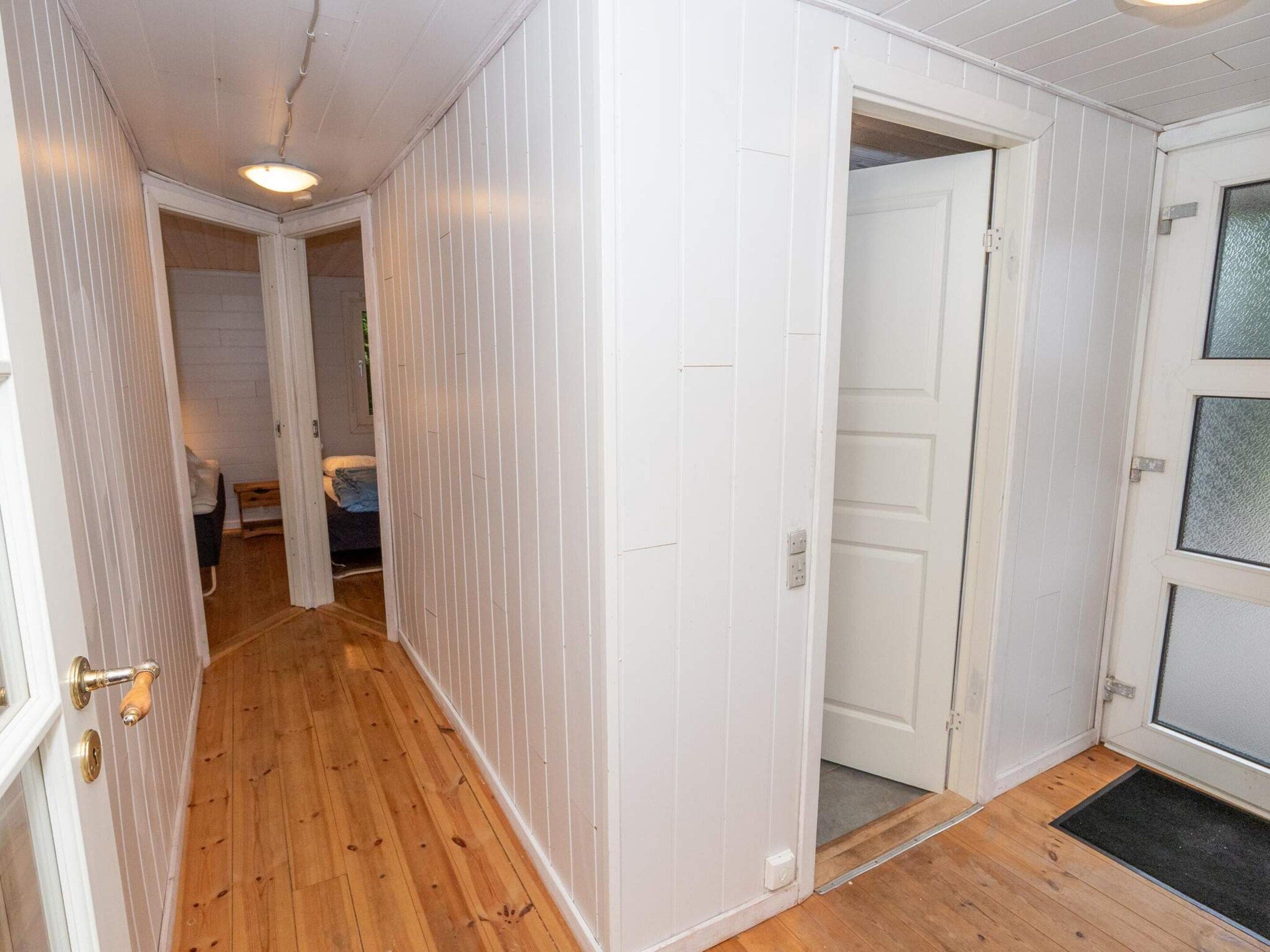 Un pasillo con suelo de madera, paredes blancas y una puerta que da a otra habitación.