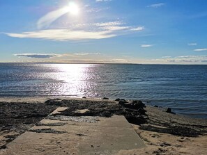 Una playa con un camino de hormigón que lleva al agua, cielo despejado y el sol reflejándose en el mar.