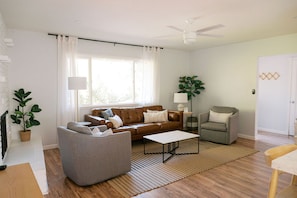 Una sala moderna con un sofá de cuero marrón, un sillón gris, una mesa de centro y una lámpara de pie. Se ve una ventana grande con cortinas blancas, una planta en maceta y piso de madera.