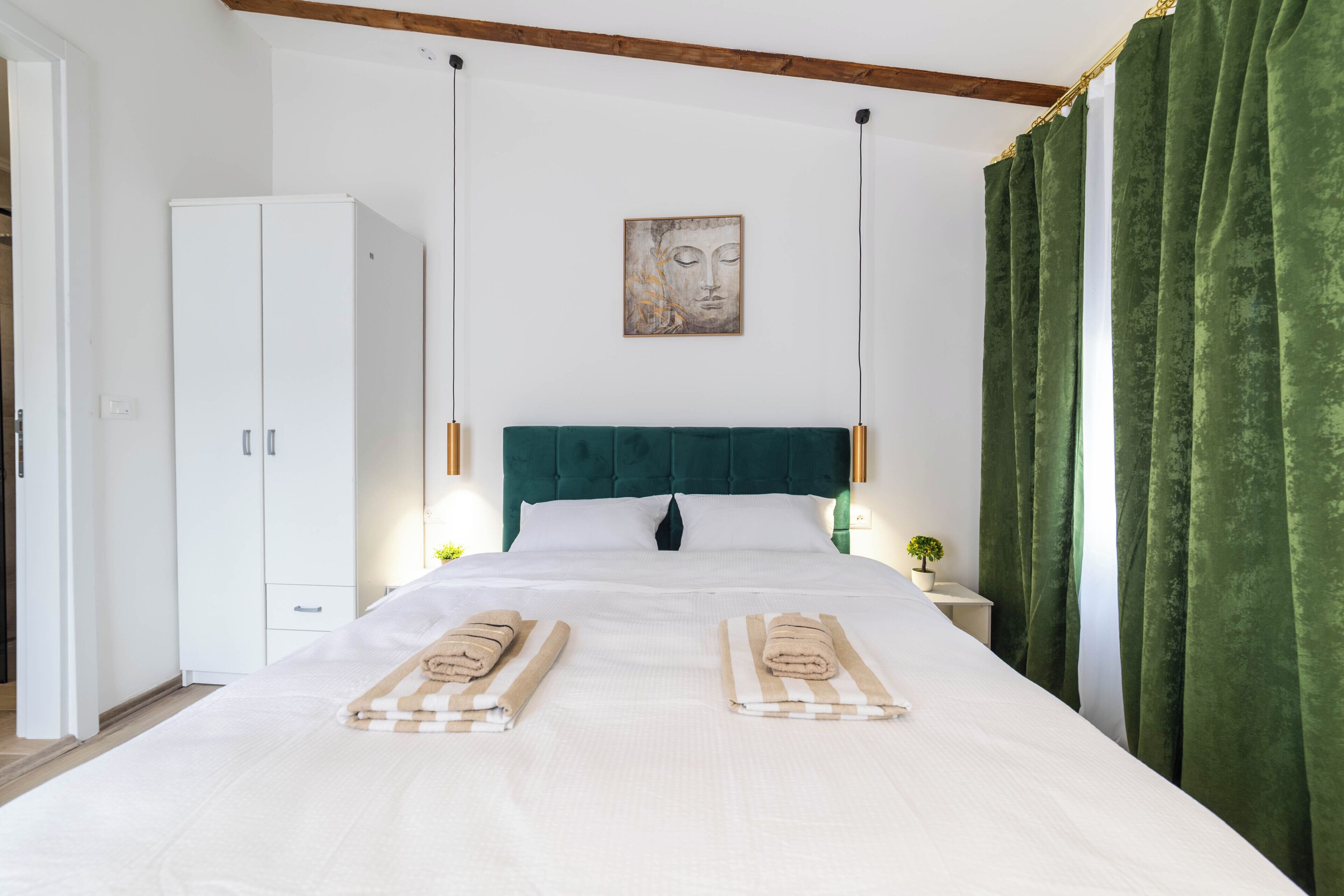Una cama bien hecha con dos toallas, un cabecero verde, paredes blancas, un armario blanco y una cortina verde.