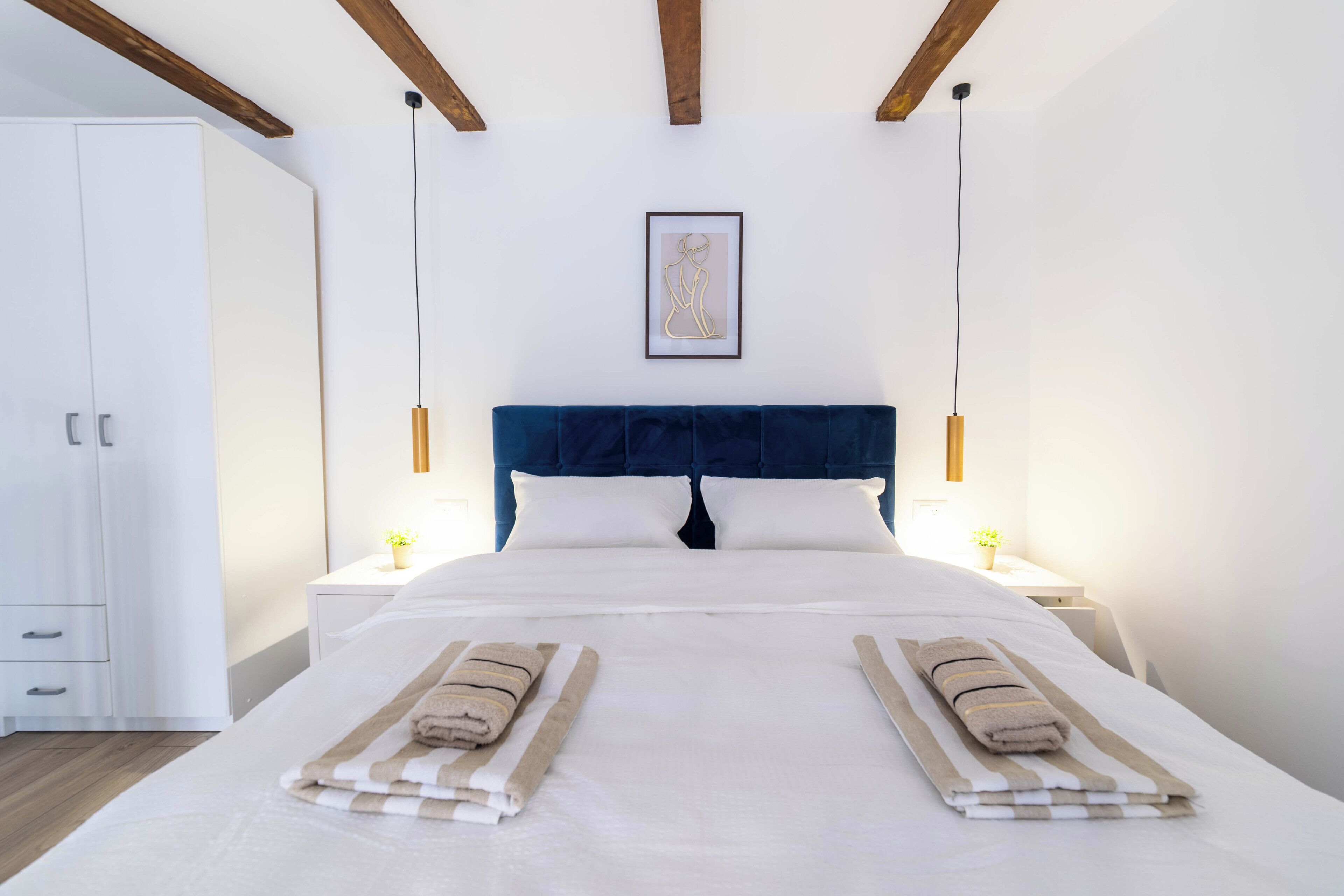 A double bed with two towels, a dark blue headboard, white walls, and a framed picture on the wall.