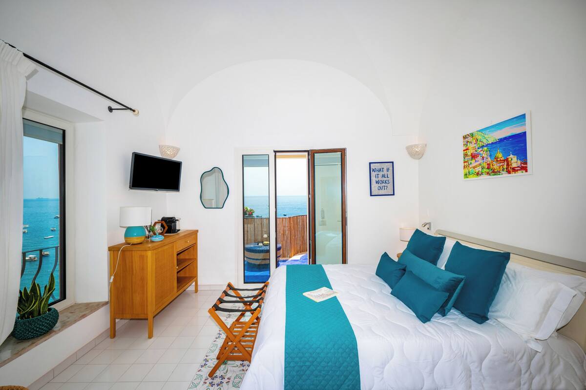 A bedroom with a bed, a desk, a TV, a window with a view of the sea, and a balcony.
