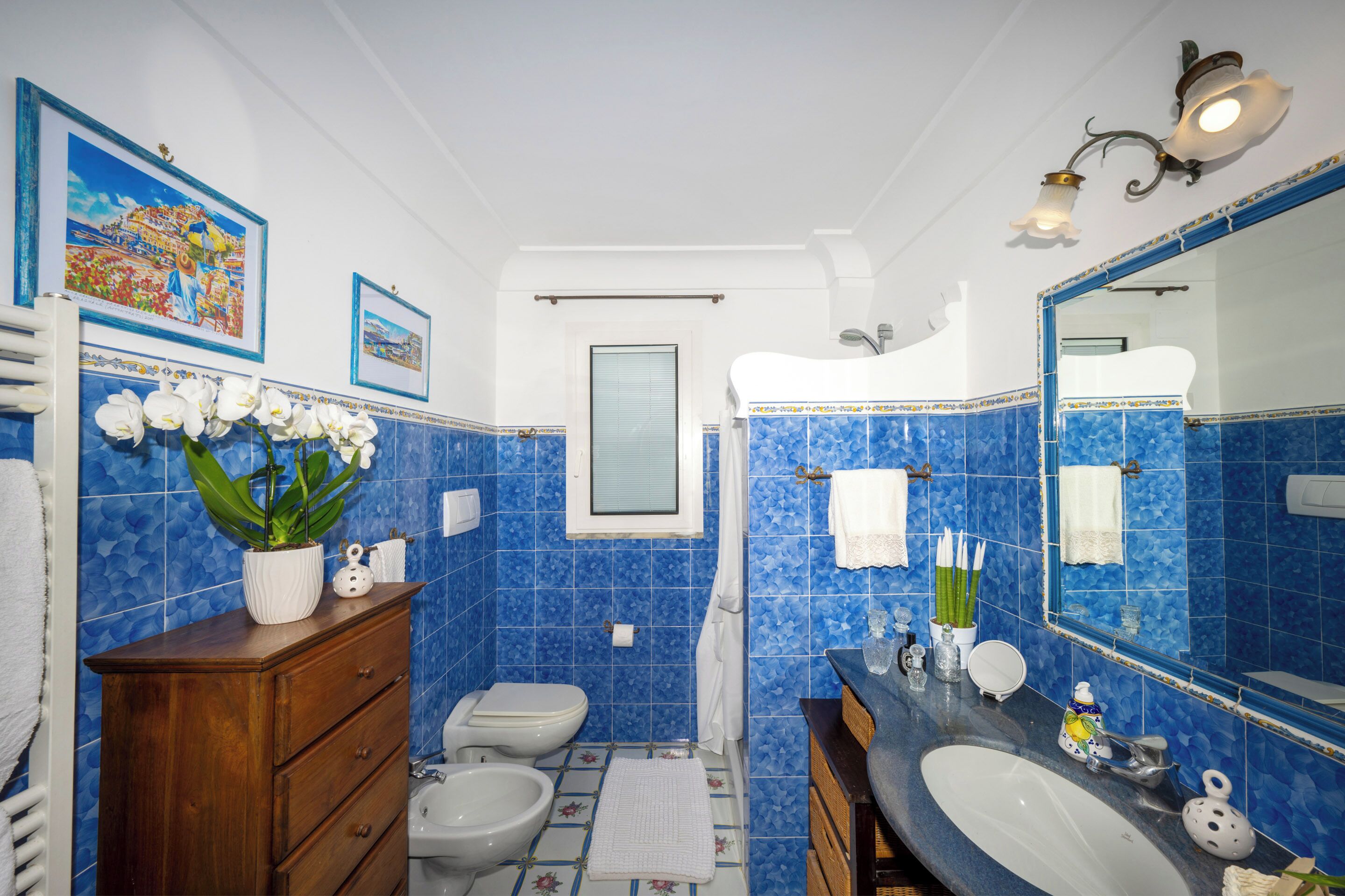 A bathroom with blue mosaic tiles, a wooden vanity, a white sink, a large mirror, and a framed picture on the wall.