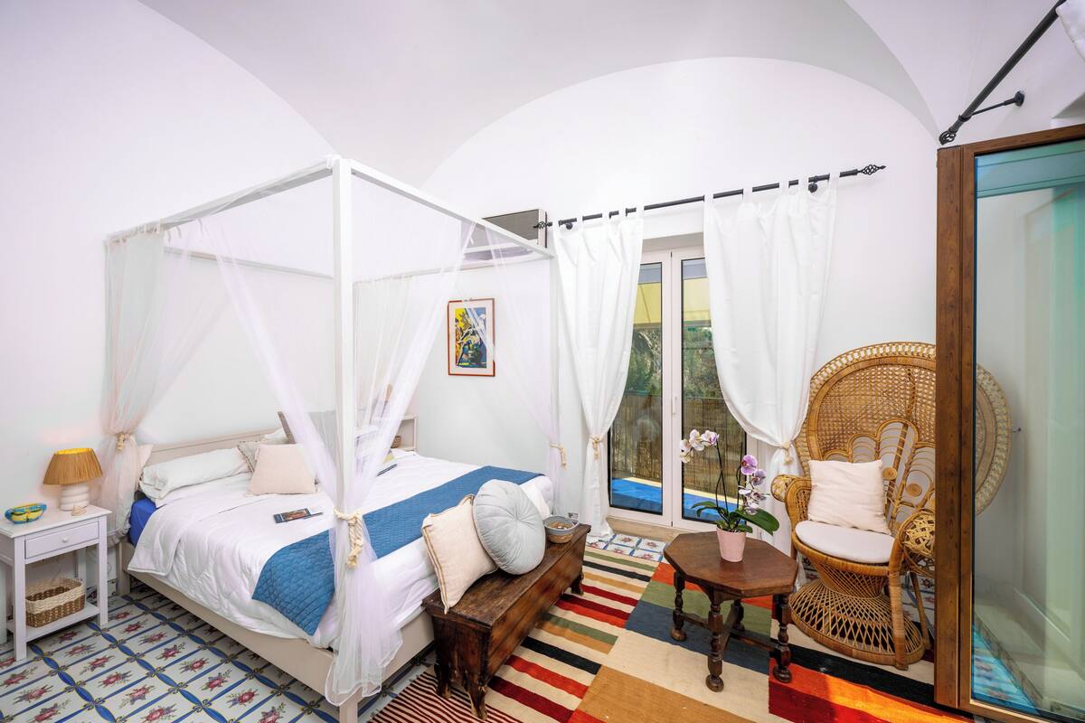 A bedroom with a four-poster bed, a wicker chair, a small table, and a view of the outdoors.
