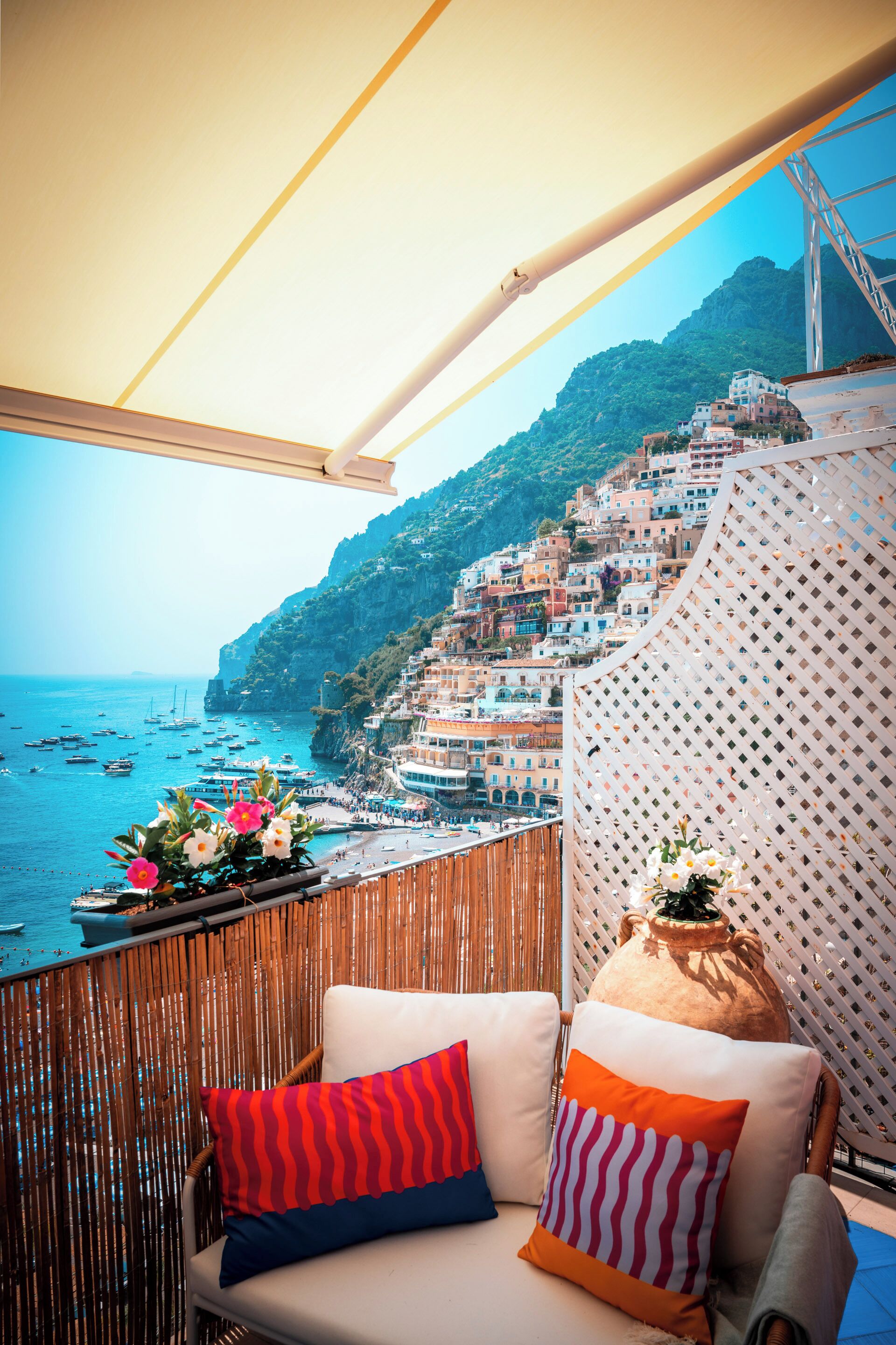 A balcony with a view of a coastal town, a wicker chair, and a large vase with flowers.