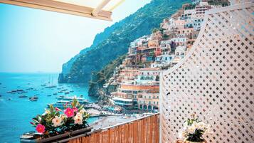 A balcony with a view of a coastal town, a wicker chair, and a large vase with flowers.