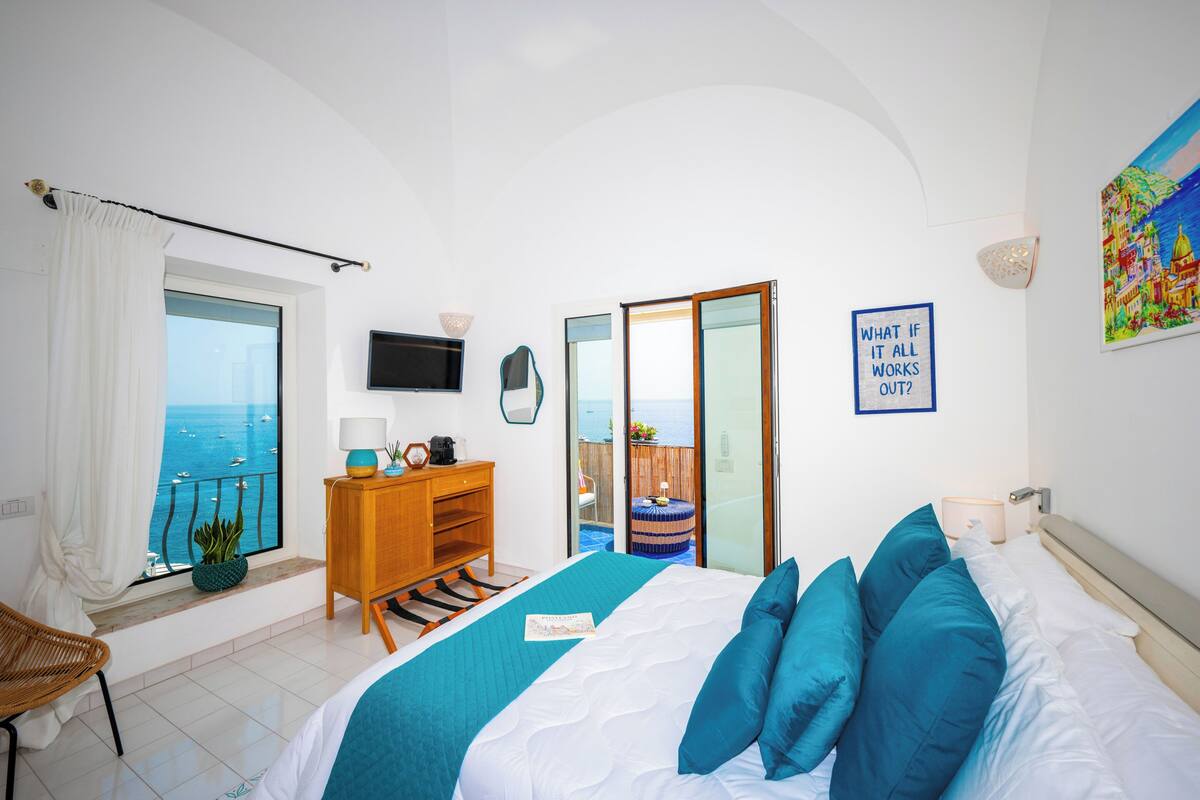 A bedroom with a bed, a desk, a chair, a TV, a window with a view of the sea, and a painting on the wall.