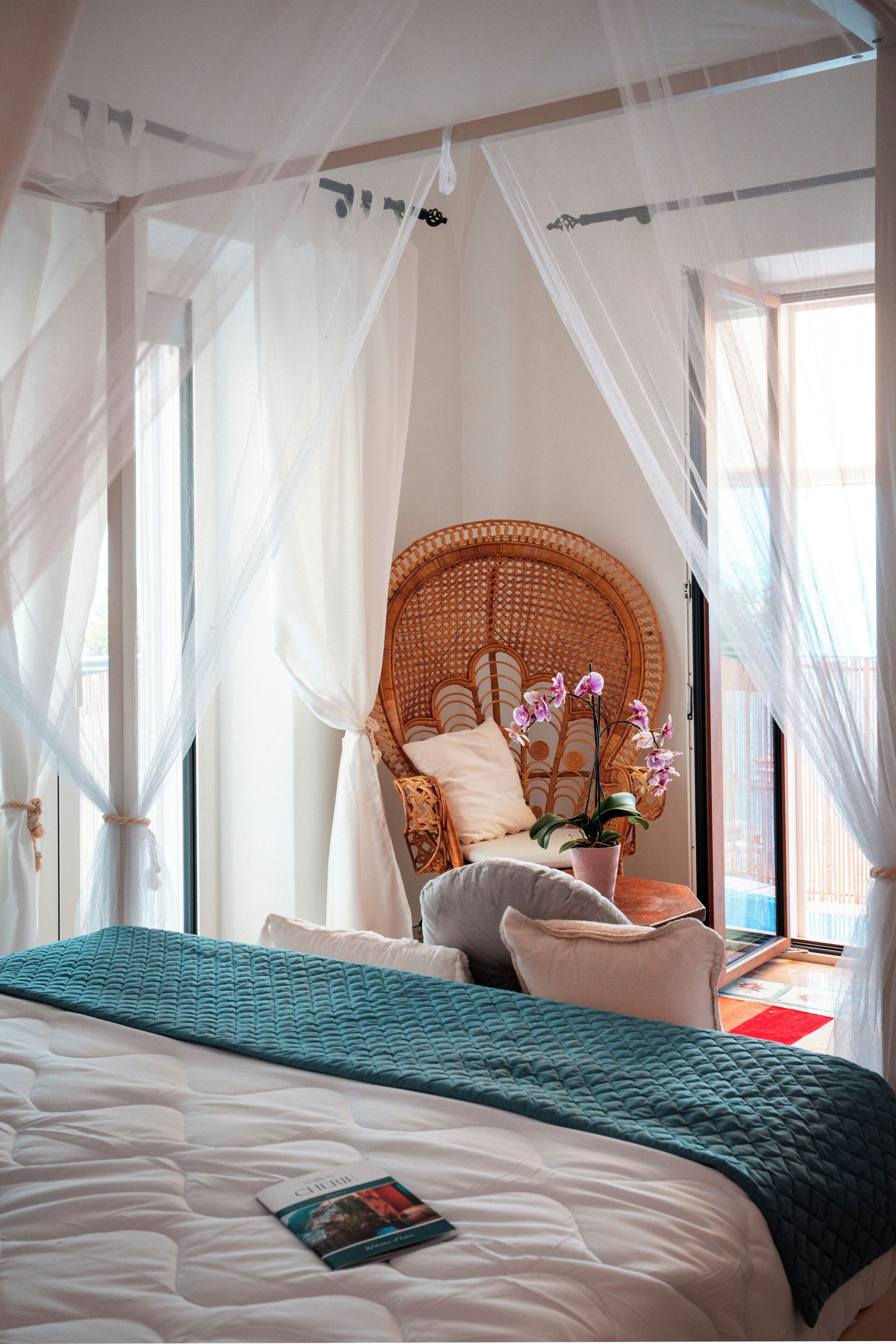 A bedroom with a bed, a wicker chair, a potted orchid, and a window with sheer curtains.