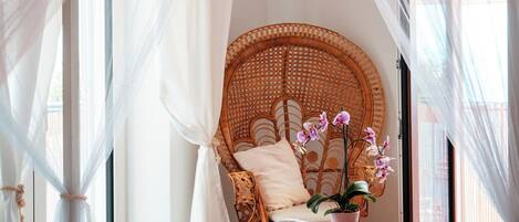 A bedroom with a bed, a wicker chair, a potted orchid, and a window with sheer curtains.