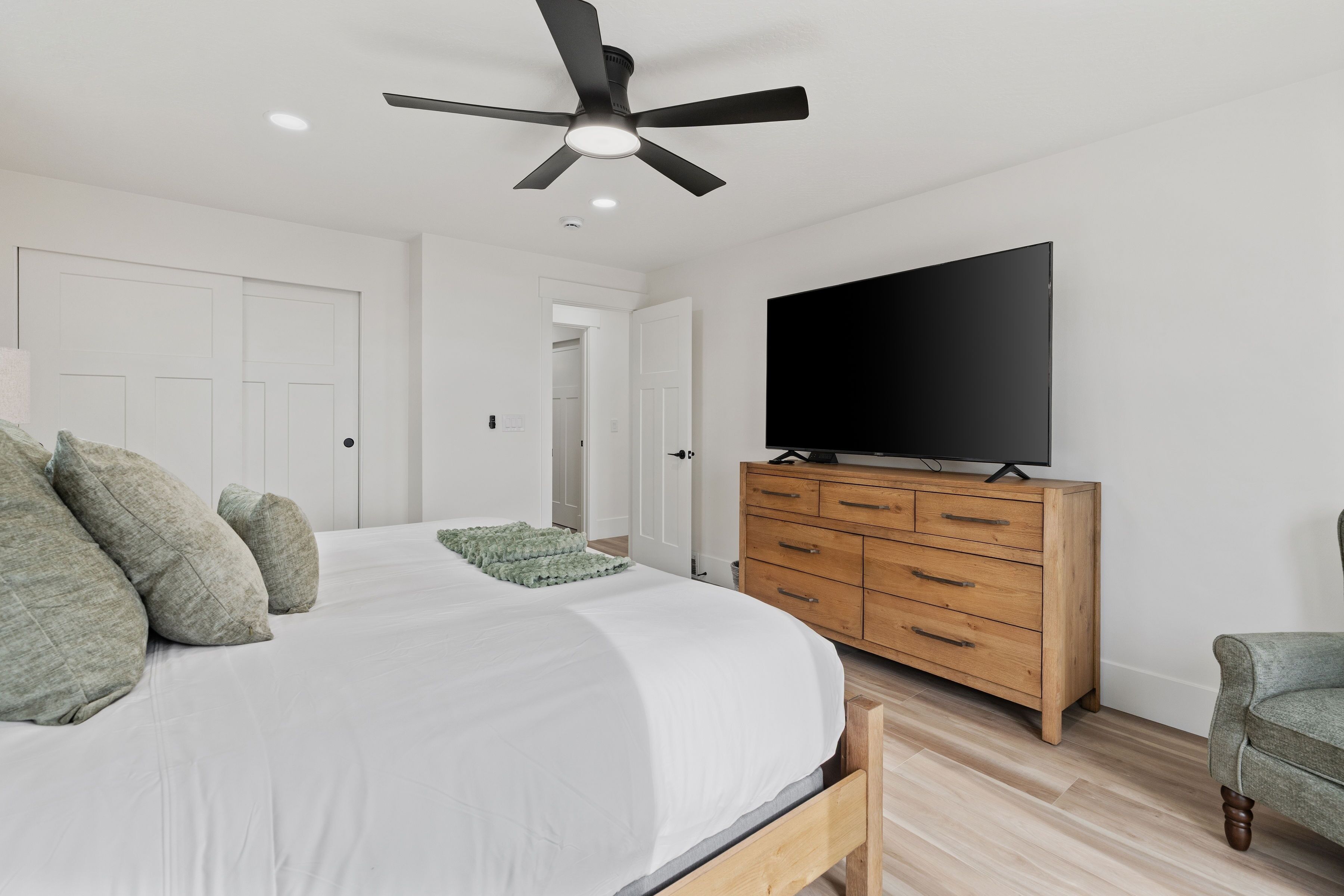 Un dormitorio con una cama, un televisor sobre un tocador de madera, un ventilador de techo y una puerta que da a otra habitación.