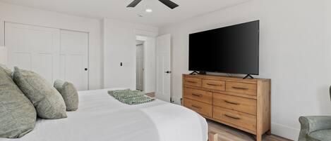 Un dormitorio con una cama, un televisor sobre un tocador de madera, un ventilador de techo y una puerta que da a otra habitación.