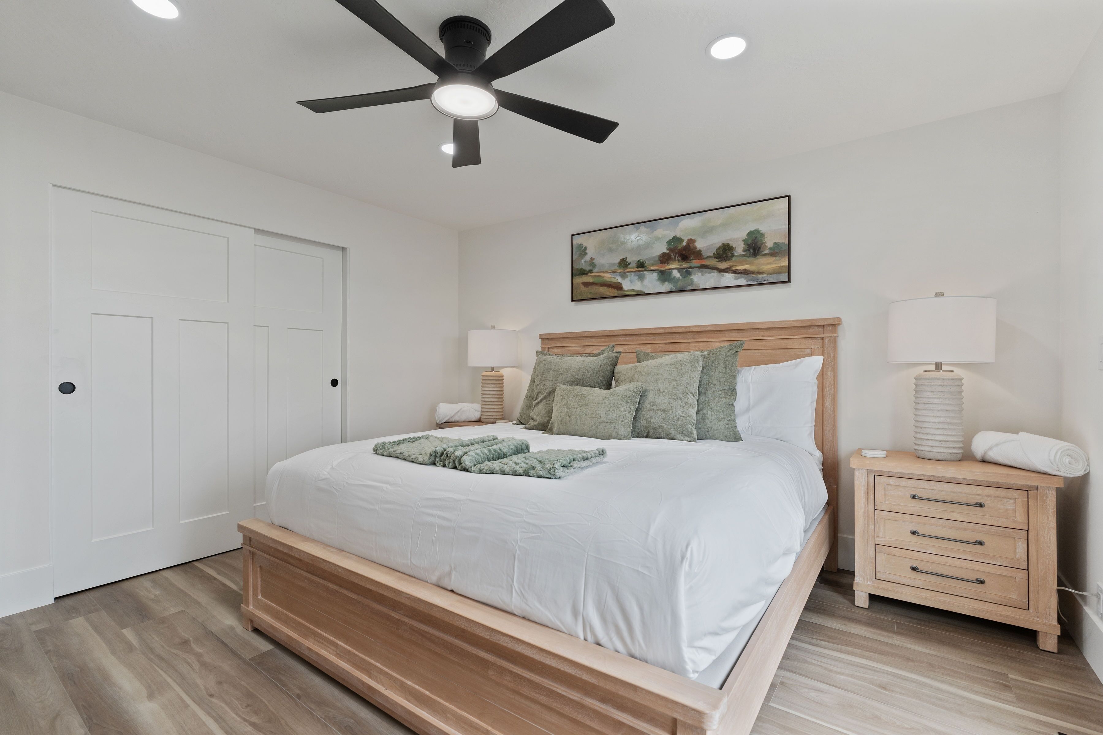 A bedroom with a bed, a nightstand, a ceiling fan, and a painting on the wall.