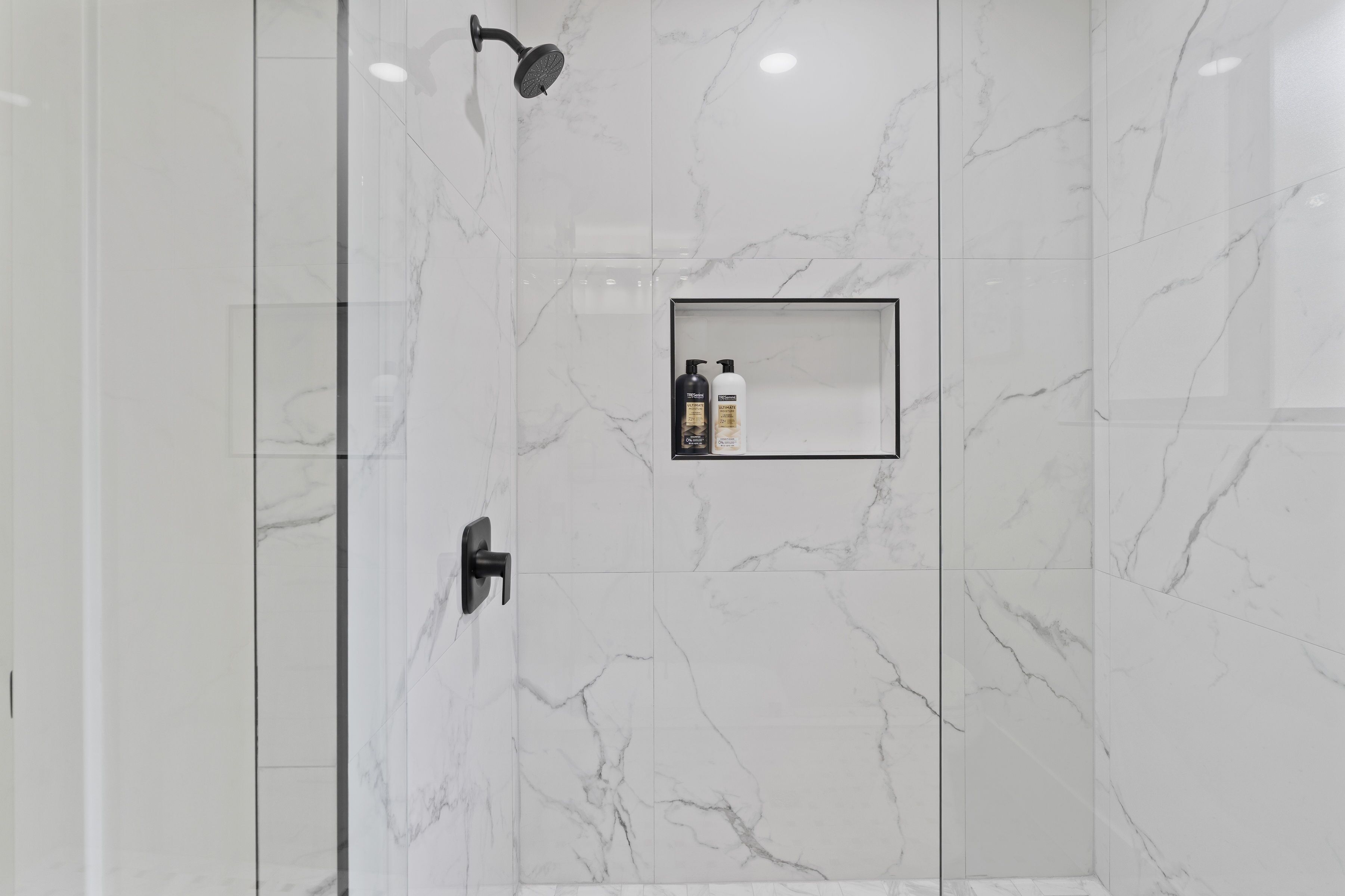 A modern bathroom with white marble tiles, a glass-enclosed shower, and a small shelf with toiletries.