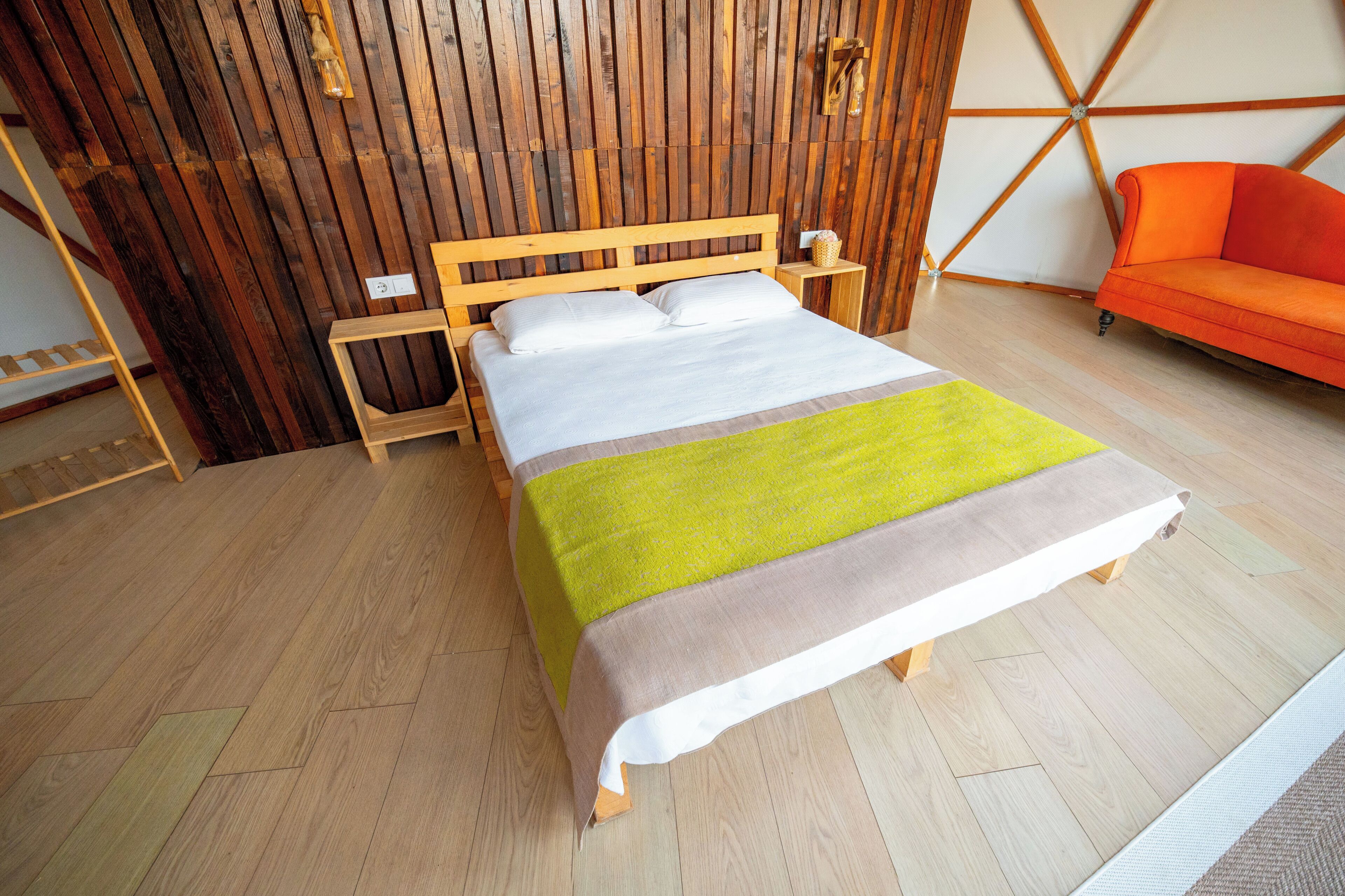 A room with a wooden bed, a wooden nightstand, a bright orange sofa, and a wooden wall with a geometric pattern.